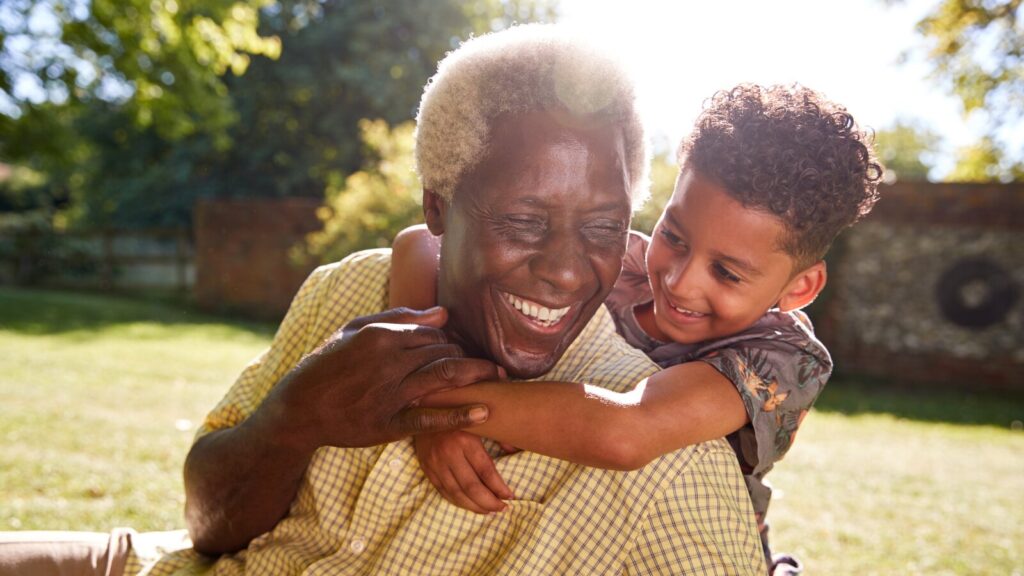 Senior Black Man Sitting On Grass, Embraced By His Grandson